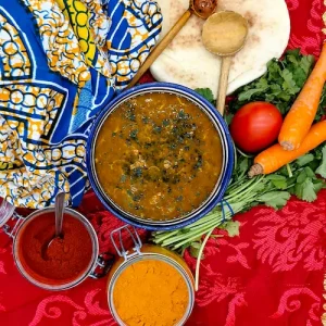 une soupe marocaine nommée chorba est posée sur une table à la décoration orientale avec les légumes et les épices que l'on retrouve dans la recette. Des cuillères en bois typiques sont posées à côté du bol.