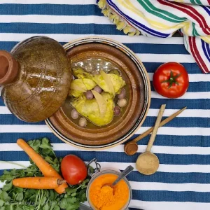 Un tajine de poulet, citron confit et olives posé sur une nappe orientale et bordé de cuillères en bois, tomate, carottes, percil.