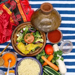 Un tajine de poulet accompagné de légumes cuits dans le plat typique en terre cuite du Maroc. La décoration de la table évoquent le Maghreb avec des tissus colorés, des légumes du soleil et des épices colorées.