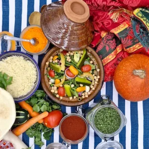 Un couvercle à tajine est posé sur le côté d'un plat en terre cuite oriental. À l'intérieur les légumes sont cuits à la vapeur et dans leur jus. Un bol de semoule accompagne le tajine. Sur la table, nous pouvons voir des légumes et des épices qui évoquent le Maroc.