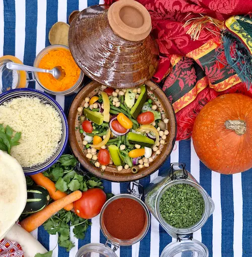 Un couvercle à tajine est posé sur le côté d'un plat en terre cuite oriental. À l'intérieur les légumes sont cuits à la vapeur et dans leur jus. Un bol de semoule accompagne le tajine. Sur la table, nous pouvons voir des légumes et des épices qui évoquent le Maroc.