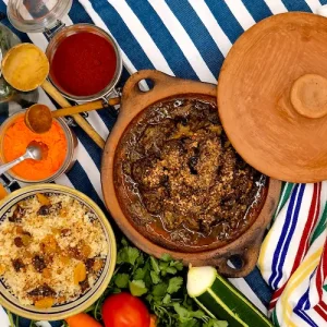 Un plat du Maroc à base de bœuf est présenté dans son plat de cuisson en terre cuite. Le plat est accompagnée d'un semoule garnie de fruits sec, raisins et abricots, le tout sur une table au décor coloré du Maghreb