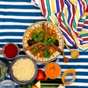 sur une nappe bleue et blanche, une serviette multicolore bleue, jaune, verte et blanche est disposée. La serviette vient entourer une généreuse assiette de couscous merguez délicieusement présentée. Sur la table, de la semoule, des épices et légumes de saison semblent présents pour accompagner le couscous.