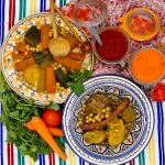 Un couscous est disposé dans une assiette aux couleurs typiques d'Afrique du nord. La semoule et les légume semblent encore chauds. Dans une autre assiette, juste à côté, la viande accompagnant le couscous royal est prête à être servie. Le plat contient, des boulettes de kefta, des merguez, du poulet et de l'agneau. Un décor oriental invite à la dégustation du plat.