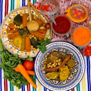 Un couscous est disposé dans une assiette aux couleurs typiques d'Afrique du nord. La semoule et les légume semblent encore chauds. Dans une autre assiette, juste à côté, la viande accompagnant le couscous royal est prête à être servie. Le plat contient, des boulettes de kefta, des merguez, du poulet et de l'agneau. Un décor oriental invite à la dégustation du plat.