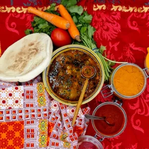 Une soupe orientale servie dans un bol typique du Maroc sur une nappe rouge aux cloueurs de l'Afrique du nord. Les épices et le pain marocains accompagnent cette soupe. Une cuillère en bois originaire des pays du Maghreb est plongée dans le contenant.