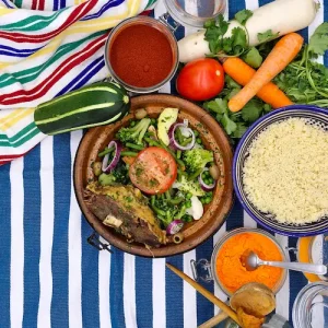 Un tajine d'agneau accompagné de ses légumes colorés sont disposés dans un plat à tajine en terre cuite. Le tout est présenté sur une table aux couleurs vives rappelant les origines orientales et maghrébines du plat.