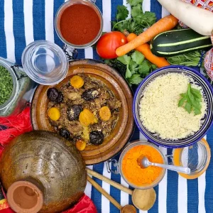 Un tajine de bœuf cuit avec ses fruits secs et parfumés. Le couvercle du plat à tajine est déposé sur le côté de l'assiette invitant à découvrir le contenu du plat. Le tout est bordé d'épices, de légumes frais, d'herbes, sur une table au décor oriental.