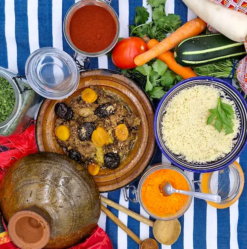 Un tajine de bœuf cuit avec ses fruits secs et parfumés. Le couvercle du plat à tajine est déposé sur le côté de l'assiette invitant à découvrir le contenu du plat. Le tout est bordé d'épices, de légumes frais, d'herbes, sur une table au décor oriental.
